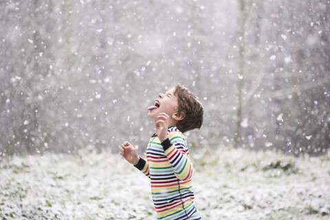 Seitenansicht eines glücklichen Jungen, der seine Zunge herausstreckt, während er auf einem Feld bei Schneefall steht, lizenzfreies Stockfoto
