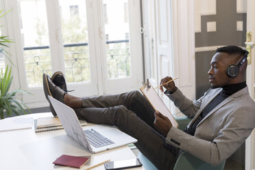 Young businessman sitting with feet up at desk in the office working and listening to music - GRSF00015