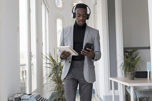 Porträt eines jungen Geschäftsmannes mit Kopfhörern und Notizbuch im Büro, der auf sein Handy schaut - GRSF00017