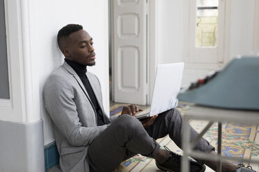 Porträt eines jungen Geschäftsmannes, der im Büro auf dem Boden sitzt und an einem Laptop arbeitet - GRSF00023