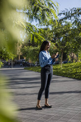 Businesswoman using smartphone, holding laptop bag in park - MAUF01852