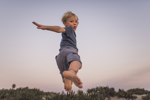 Niedriger Blickwinkel des Jungen springen auf Sand am Strand gegen den Himmel bei Sonnenuntergang, lizenzfreies Stockfoto