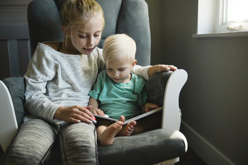 Schwester und Bruder schauen sich ein Buch an, während sie auf einem Stuhl zu Hause sitzen - CAVF58475