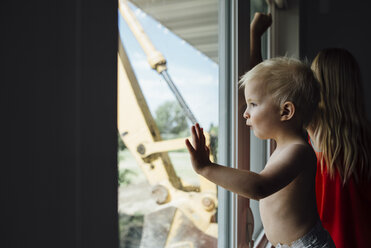 Seitenansicht eines hemdsärmeligen kleinen Jungen, der durch das Fenster schaut, während er bei seiner Schwester zu Hause steht - CAVF58494