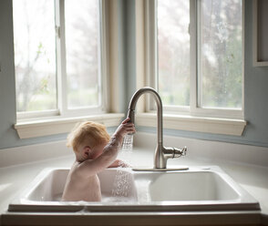Niedlicher kleiner Junge, der den Wasserhahn hält, während er in der Küchenspüle vor dem Fenster sitzt - CAVF58518