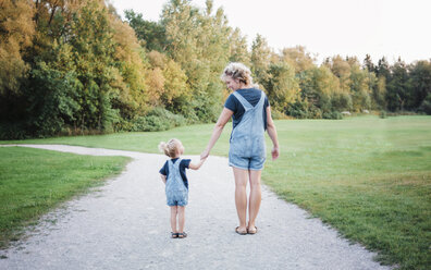 Rear view of mother and daughter wearing bib overalls while holding hands on dirt road - CAVF58545