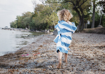 Rückansicht eines in ein Handtuch eingewickelten Mädchens beim Spaziergang am Seeufer - CAVF58550