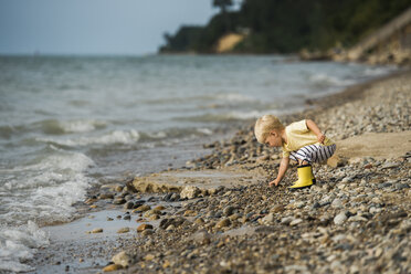 Side view of boy picking pebbles while playing on shore - CAVF58559