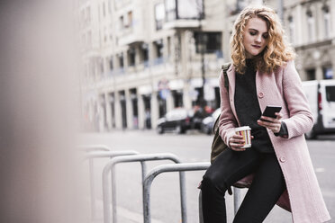 Geschäftsfrau, die ein Smartphone benutzt, während sie auf einem Geländer in der Stadt sitzt - CAVF58575