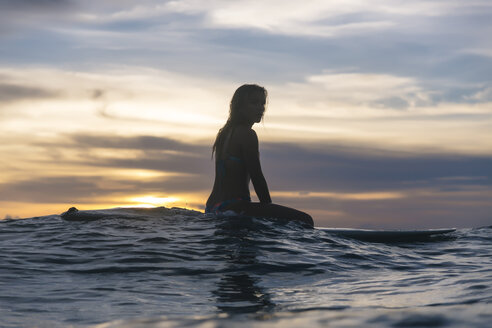 Seitenansicht einer jungen Frau, die auf einem Surfbrett im Meer sitzt, gegen einen bewölkten Himmel bei Sonnenuntergang - CAVF58685