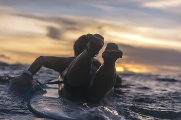 Rückansicht einer jungen Frau, die auf einem Surfbrett liegt, während sie bei Sonnenuntergang im Meer schwimmt - CAVF58686