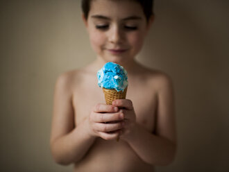 Shirtless boy holding ice cream while standing against wall at home - CAVF58692