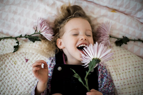 Draufsicht auf ein fröhliches Mädchen mit Gerbera, das zu Hause auf dem Bett liegt - CAVF58718