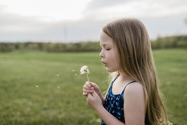 Side view of girl blowing dandelion flower on grassy field - CAVF58723