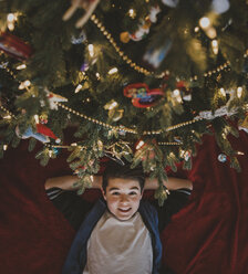 Overhead portrait of boy with hands behind head lying by Christmas tree at home - CAVF58773
