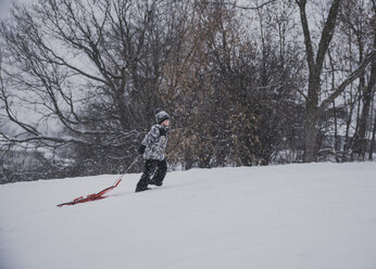Seitenansicht eines Jungen, der einen Schlitten zieht, während er auf einem verschneiten Feld gegen kahle Bäume läuft - CAVF58775