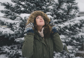 Frau mit Pelzmantel, die bei Schneefall gegen Bäume blickt - CAVF58779