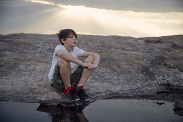 Boy looking away while sitting by stream on Arabia Mountain against cloudy sky - CAVF58790