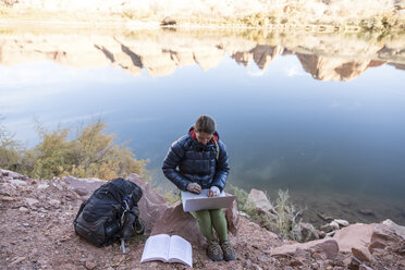 Hochformatige Ansicht einer Wanderin, die einen Laptop benutzt, während sie auf einem Felsen am See sitzt - CAVF58822