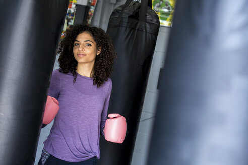 Porträt einer weiblichen Boxerin mit rosa Boxhandschuhen, die an einem Boxsack im Fitnessstudio steht - CAVF58836