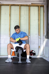 Male boxer wearing boxing gloves while sitting in gym - CAVF58842