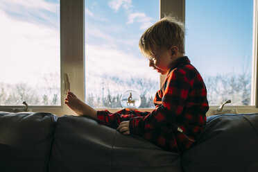 Side view of boy looking at decor while sitting on sofa by window at home - CAVF58853