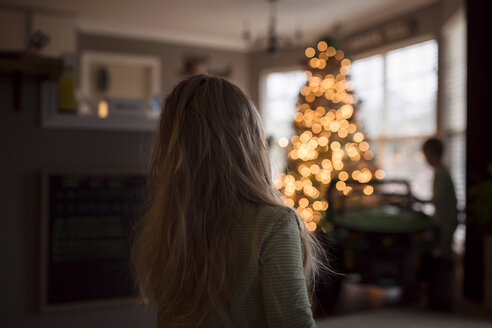 Rückansicht der Schwester, die zu Hause steht, während der Bruder am beleuchteten Weihnachtsbaum sitzt - CAVF58874