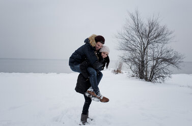 Glückliche Frau, die einen männlichen Freund huckepack nimmt, während sie am schneebedeckten Strand vor dem Himmel steht - CAVF58895