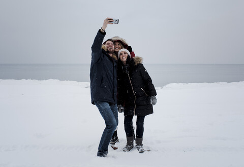 Freunde, die ein Selfie machen, während sie am schneebedeckten Strand vor dem Himmel stehen, lizenzfreies Stockfoto