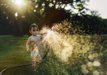 Shirtless baby girl spraying water with garden hose at yard - CAVF58936