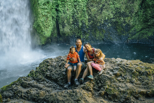 Porträt einer glücklichen Familie, die auf Felsen vor einem Wasserfall im Wald sitzt - CAVF58974