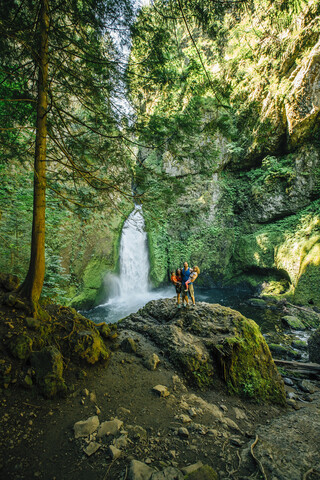 Familie steht auf Felsen vor einem Wasserfall im Wald, lizenzfreies Stockfoto