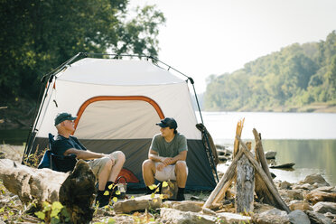 Friends talking while relaxing at campsite by lake against sky - CAVF59008