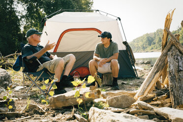 Männliche Freunde unterhalten sich beim Sitzen auf dem Campingplatz am See - CAVF59011