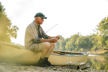 Älterer Mann, der auf einem Boot am Seeufer sitzend seine Angelausrüstung gegen den klaren Himmel ausrichtet - CAVF59023