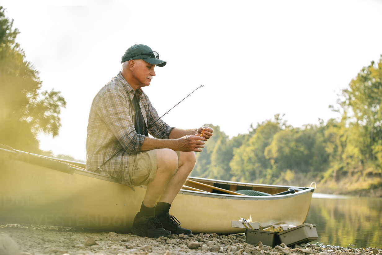 Senior man adjusting fishing tackle while sitting on boat at lakeshore  against clear sky stock photo