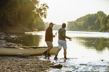 Male friends holding fishing rods talking while walking at lakeshore - CAVF59036