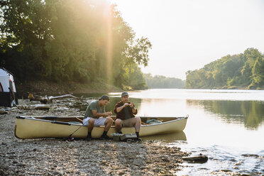 Male friends adjusting fishing tackles while sitting on boat at campsite by lake - CAVF59038