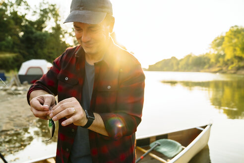 Mid adult man tying tackle to fishing line while standing at lakeshore - CAVF59041