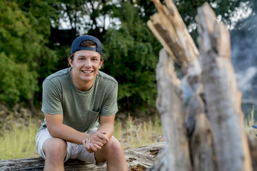 Portrait of smiling young man sitting on log at campsite during sunset - CAVF59051