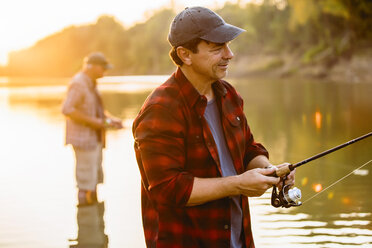 Man fishing while friend standing in background during sunset - CAVF59059