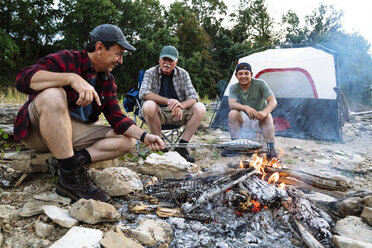 Mittlerer Erwachsener, der beim Zelten mit Freunden während des Sonnenuntergangs Fisch am Lagerfeuer kocht - CAVF59066