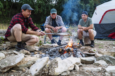 Mittlerer Erwachsener, der Fisch auf dem Lagerfeuer kocht, während er mit Freunden auf dem Campingplatz bei Sonnenuntergang sitzt - CAVF59068