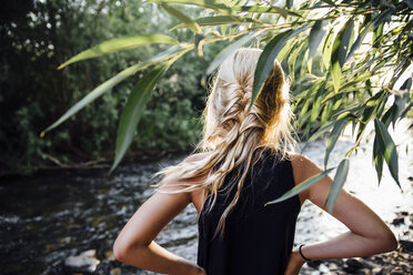 Rückansicht eines unbekümmerten Mädchens mit Händen auf der Hüfte, das am Fluss im Wald steht - CAVF59109