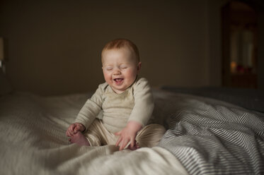 Cheerful baby boy with eyes closed sitting on bed - CAVF59167