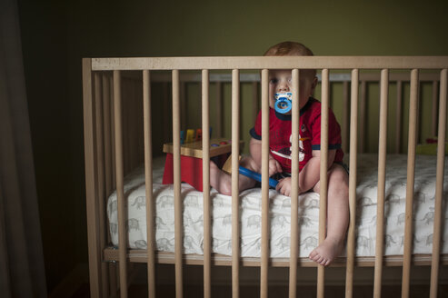 Porträt eines kleinen Jungen, der am Schnuller nuckelt, während er zu Hause in seinem Kinderbett sitzt - CAVF59170