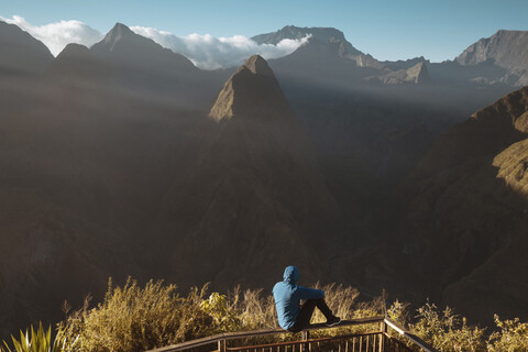 Rückansicht eines Mannes, der auf einem Aussichtspunkt sitzend die Berge betrachtet, lizenzfreies Stockfoto