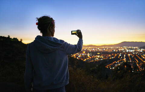 Chile, Santiago, Frau nimmt Handy-Foto in den Bergen über der Stadt bei Sonnenuntergang, lizenzfreies Stockfoto