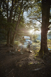 Chile, Santa Magda, Rio Maniguales, boy sitting at campfire in forest with camper van in background - SSCF00223