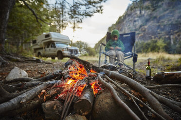 Chile, Santa Magda, Rio Maniguales, Junge sitzt am Lagerfeuer im Wald mit Wohnmobil im Hintergrund - SSCF00224